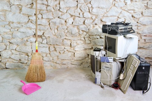 The image shows a corner of an indoor or garage space with a beige, textured brick wall in the background. In the foreground, a pink plastic dustpan and a small straw broom with a wooden handle and natural bristles are positioned on the light grey concrete floor, indicating cleaning tools. To the right, a collection of old electronic equipment, including a computer monitor, desktop tower, and various cables, is stacked against the wall. An old wooden or plastic garden rake with a yellow handle is leaning against the wall on the left side of the image. The setting appears to be a storage or utility area, typical for disposing of or organising waste and unwanted household items, consistent with services offered by Commercial Waste Removal Hendon in the Hendon area. The cluttered arrangement and the presence of waste-related items highlight the need for professional rubbish removal in a domestic or commercial context within the local London postcode area.
