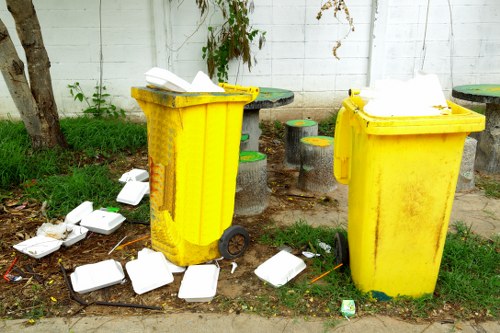 Two yellow wheeled rubbish bins, made of hard plastic with textured surfaces, are positioned on a pavement outside a white brick wall. The bin on the left has its lid partially open, revealing some scattered white foam containers beside and around it, with additional containers on the ground nearby. The bin on the right appears closed with foam containers stacked on top of its partially open lid. In the background, some small wooden stools or plant stands with green and yellow finishes are visible, along with patches of grass and small shrubs growing near the wall. The scene suggests an outdoor waste collection area, consistent with commercial rubbish removal services in Hendon, London, and indicates a recent disposal of food packaging or delivery containers.