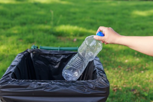 A person's hand is seen placing a clear plastic water bottle into a black plastic rubbish bin outdoors. The bottle is partially crushed, with a blue cap, and the bin is situated on a paved surface with blurred green grass in the background, indicating a garden or park setting. The scene reflects a typical waste disposal action, relevant to waste management and rubbish removal services offered by companies like Commercial Waste Removal Hendon, which operates in the Hendon area. The lighting appears natural, suggesting daylight, and the overall environment is clean and orderly, with emphasis on recycling and waste separation practices commonly associated with commercial and residential waste collection in London suburbs. The image underscores the importance of responsible waste disposal in local communities such as Hendon, aligned with professional rubbish removal services.