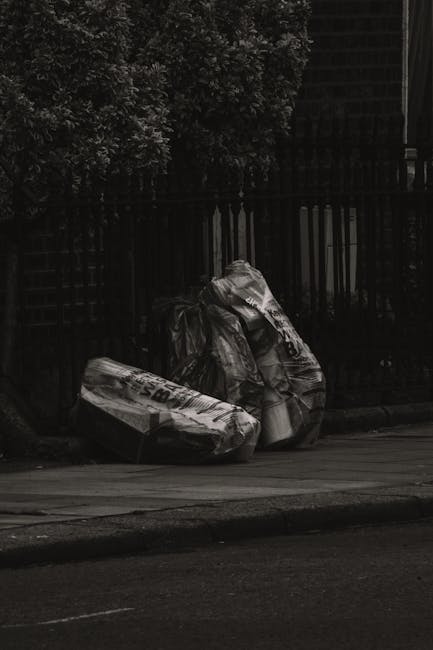 Two large black plastic garbage bags filled with waste sit on the pavement next to a black metal fence, which runs parallel to the adjacent road. The bags are slightly crumpled and appear to contain m